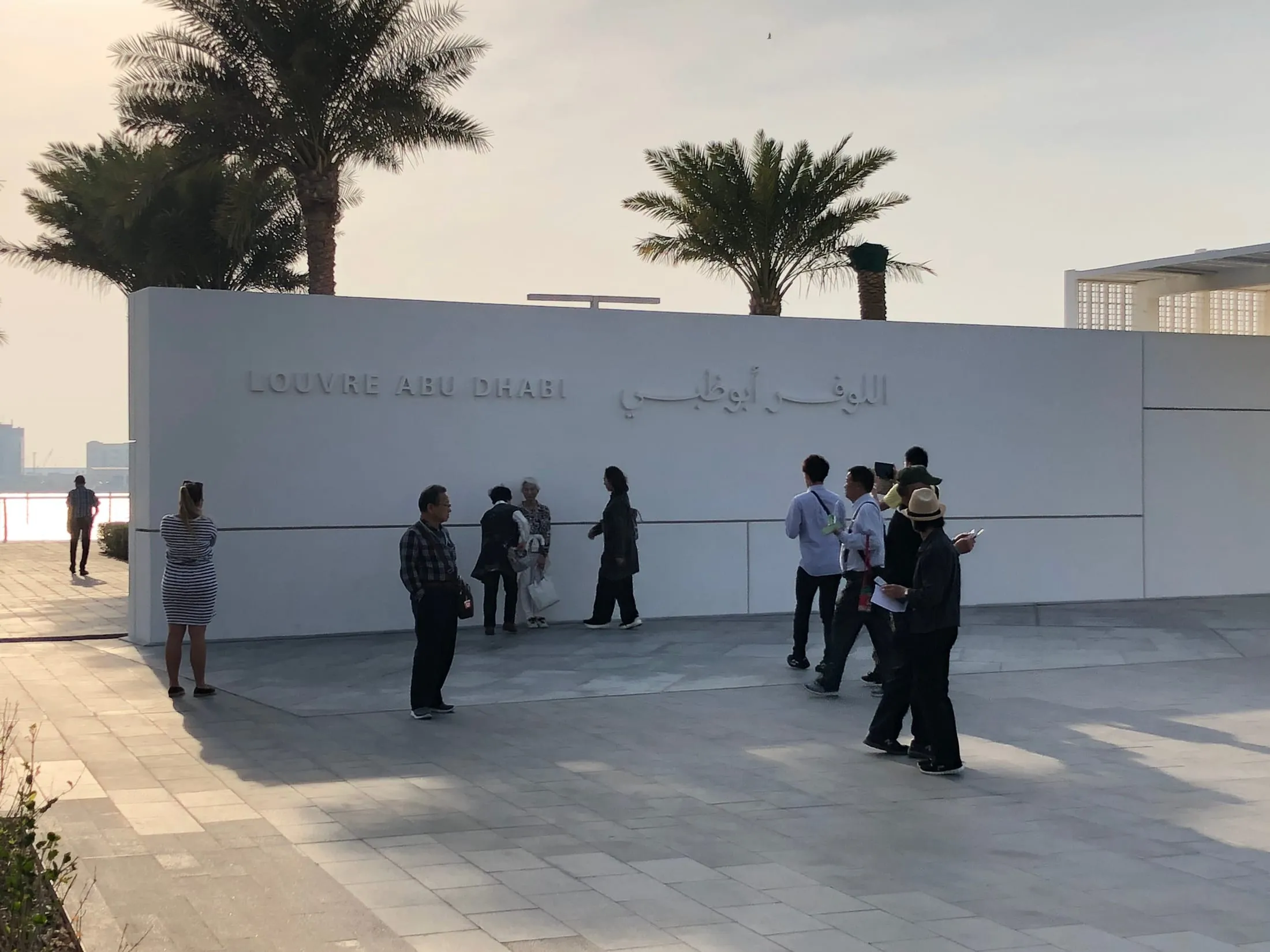 Вход в Лувр Абу-Даби: стена с надписью «Louvre Abu Dhabi» на английском и арабском, пальмы на фоне